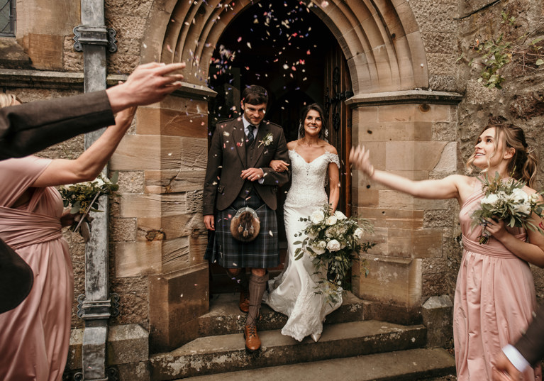 groom and bride walking out of church doorway with bridesmaids throwing confetti